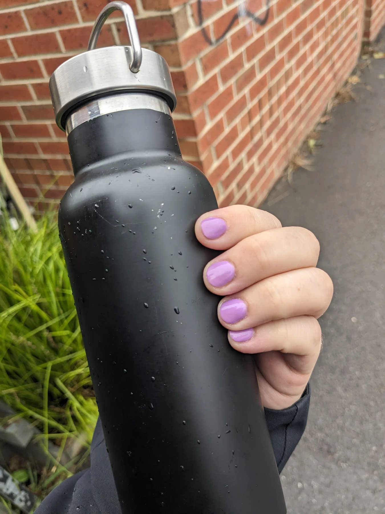 Lilac nails gripping a black water bottle against brick wall